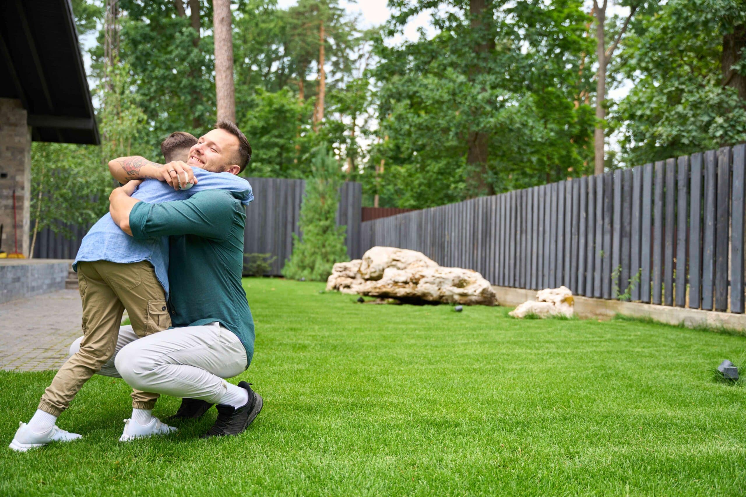 Father and son hugging on healthy green lawn with trees and fence in background