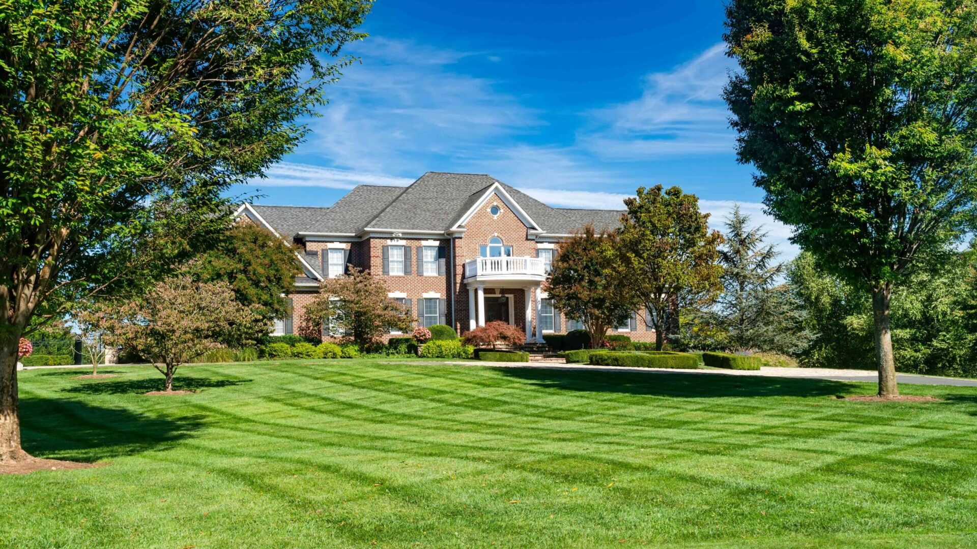 Well-maintained green ryegrass lawn in front of brick home with mature trees and clear blue sky